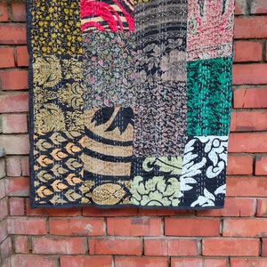 May include: A patchwork quilt with a variety of colorful fabrics, including black, gold, red, green, and brown. The quilt is made of squares and rectangles of different sizes and patterns. The quilt is hanging on a brick wall.