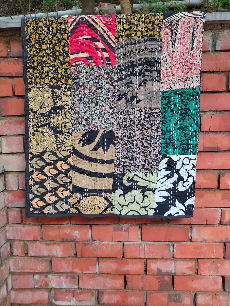May include: A patchwork quilt with a variety of colorful fabrics, including black, gold, red, green, and brown. The quilt is made of squares and rectangles of different sizes and patterns. The quilt is hanging on a brick wall.
