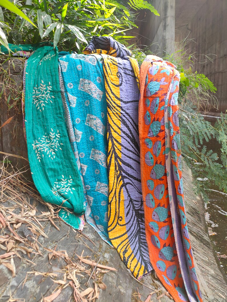 May include: Four colorful kantha quilts with different patterns, including a teal quilt with white floral designs, a blue quilt with white geometric designs, a yellow and purple quilt with black swirls, and an orange quilt with blue fish designs.