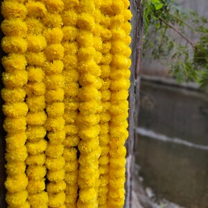 May include: A garland of bright yellow marigold flowers, strung together in a long strand. The garland is hanging from a wooden surface.