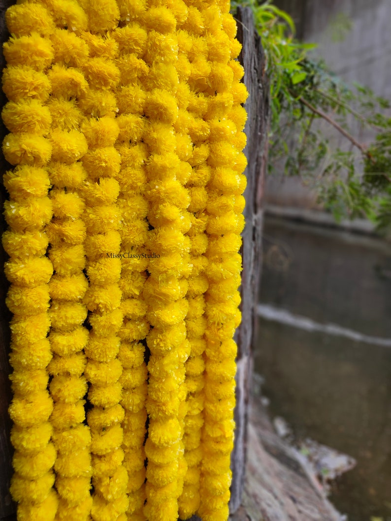 May include: A garland of bright yellow marigold flowers, strung together in a long strand. The garland is hanging from a wooden surface.