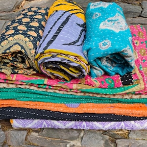 Könnte beinhalten: Ein Stapel bunter, gemusterter Quilts und Decken. Die gefalteten Decken zeigen verschiedene Designs, darunter florale, geometrische und abstrakte Muster in Blau-, Gelb-, Rosa- und Grüntönen. Die Quilts sind auf einer Steinoberfläche gestapelt.