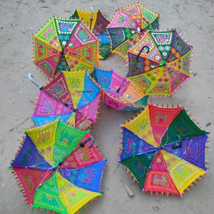 Könnte beinhalten: Eine Sammlung farbenfroher, offener Regenschirme mit aufwendigen Stickereien. Die Regenschirme weisen eine Vielzahl leuchtender Farben auf, darunter Rosa, Gelb, Grün und Blau, mit detaillierten Mustern und dekorativen Kanten. Die Griffe sind weiß oder schwarz.