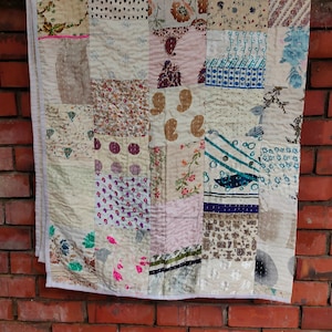 May include: A patchwork quilt made from various fabrics in shades of white, brown, pink, and blue. The quilt is made up of squares of different patterns and textures, including floral prints, paisley designs, and geometric patterns. The quilt is hanging on a brick wall.