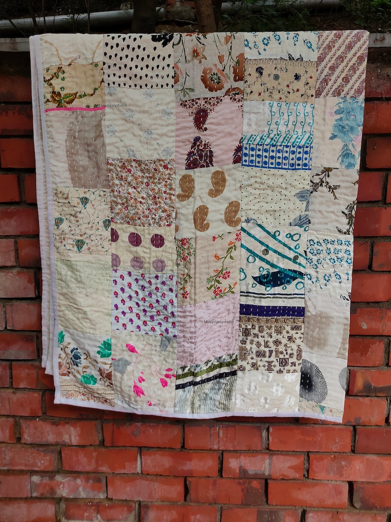 May include: A patchwork quilt made from various fabrics in shades of white, brown, pink, and blue. The quilt is made up of squares of different patterns and textures, including floral prints, paisley designs, and geometric patterns. The quilt is hanging on a brick wall.