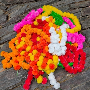 Könnte beinhalten: Eine farbenfrohe Blumengirlande mit rosa, orangefarbenen, gelben, roten, grünen und weißen Blüten. Die Girlande liegt auf einer strukturierten, grauen Steinoberfläche und zeigt eine Vielzahl floraler Elemente.