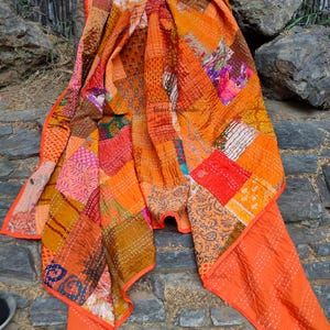Puede incluir: Un edredón de retazos en tonos naranja, rojo y marrón. El edredón está hecho de varios cuadrados de tela con diferentes patrones y texturas. Los bordes están acabados con un borde naranja liso. El edredón está drapeado sobre una superficie de piedra.