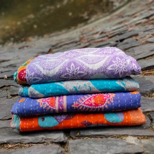 May include: A stack of four kantha quilts in various colors and patterns. The top quilt is purple with a white floral design, the second is turquoise with a white geometric design, the third is blue with a pink paisley design, and the bottom quilt is orange with a blue floral design.