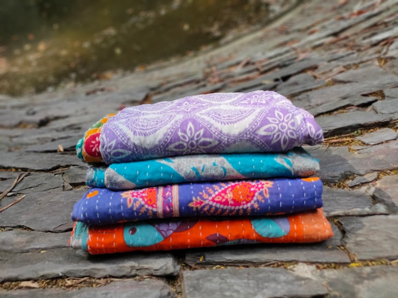 May include: A stack of four kantha quilts in various colors and patterns. The top quilt is purple with a white floral design, the second is turquoise with a white geometric design, the third is blue with a pink paisley design, and the bottom quilt is orange with a blue floral design.
