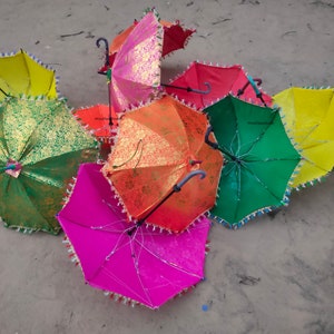 May include: A group of colorful umbrellas with gold accents and fringe trim. The umbrellas are arranged in a circle on a sandy surface.