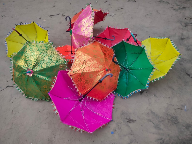 May include: A group of colorful umbrellas with gold accents and fringe trim. The umbrellas are arranged in a circle on a sandy surface.
