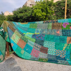 Peut inclure: Une courtepointe patchwork dans les tons de vert, sarcelle et bleu, avec une vari&eacute;t&eacute; de carr&eacute;s &agrave; motifs. La courtepointe est drap&eacute;e sur une structure en bois &agrave; l'ext&eacute;rieur, avec le texte "Missy Classy Studio" visible. Les bords de la courtepointe sont verts unis.