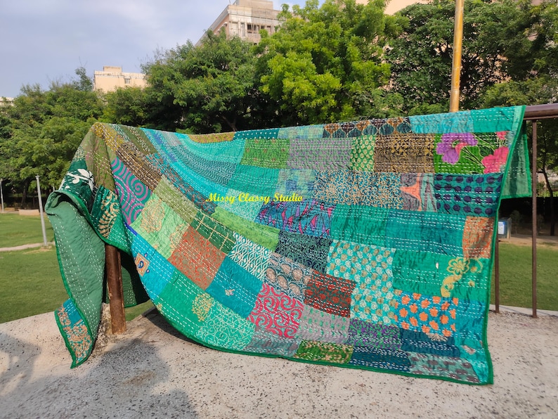 Peut inclure: Une courtepointe patchwork dans les tons de vert, sarcelle et bleu, avec une vari&eacute;t&eacute; de carr&eacute;s &agrave; motifs. La courtepointe est drap&eacute;e sur une structure en bois &agrave; l'ext&eacute;rieur, avec le texte "Missy Classy Studio" visible. Les bords de la courtepointe sont verts unis.