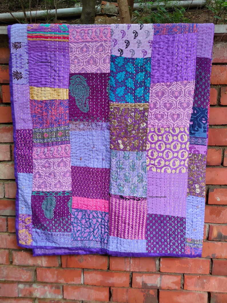 May include: A purple and blue patchwork quilt with a variety of patterns. The quilt is made of many different fabrics and is stitched together with a running stitch. The quilt is hanging on a brick wall.