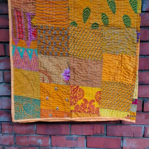 Könnte beinhalten: Eine Patchwork-Decke aus verschiedenen Stoffen in Gelb-, Orange- und Brauntönen. Die Decke besteht aus Quadraten und Rechtecken, jedes mit einem anderen Muster. Die Decke hängt an einer Backsteinwand.