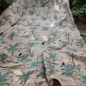 K&ouml;nnte beinhalten: Eine beige Kantha-Steppdecke mit einem sich wiederholenden Muster aus Zebras, Tukanen und Palmen. Die Steppdecke ist in der H&auml;lfte gefaltet und hat einen roten gestickten Rand.