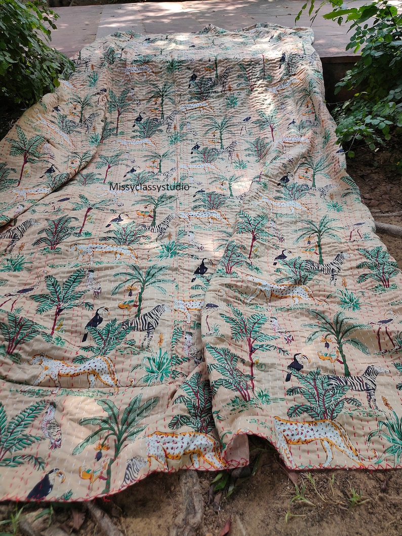 K&ouml;nnte beinhalten: Eine beige Kantha-Steppdecke mit einem sich wiederholenden Muster aus Zebras, Tukanen und Palmen. Die Steppdecke ist in der H&auml;lfte gefaltet und hat einen roten gestickten Rand.