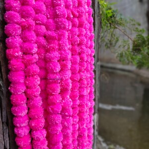 May include: A garland of bright pink paper flowers, strung together in a long strand. The garland is hanging on a wooden surface, with a blurry background of water and greenery.