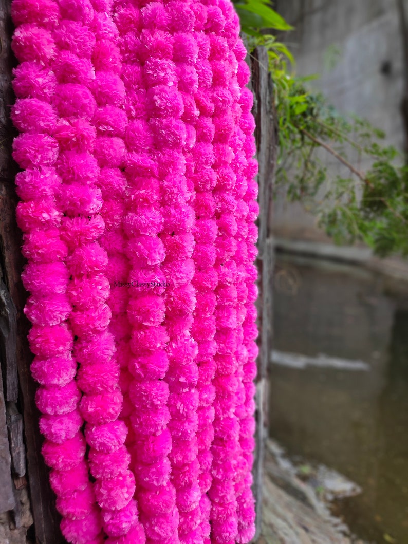 May include: A garland of bright pink paper flowers, strung together in a long strand. The garland is hanging on a wooden surface, with a blurry background of water and greenery.