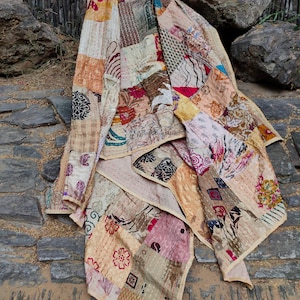 Könnte beinhalten: Ein Patchwork-Quilt mit verschiedenen Stoffmustern und -farben, darunter florale, geometrische und abstrakte Designs. Der Quilt besteht aus verschiedenen Beige-, Rosa- und Brauntönen. Der Quilt ist über einer Steinoberfläche drapiert.