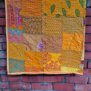 May include: A patchwork quilt made of various fabrics in shades of yellow, orange, and brown. The quilt is made up of squares and rectangles, each with a different pattern. The quilt is hanging on a brick wall.