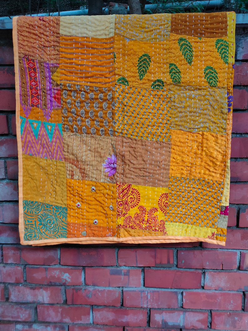 May include: A patchwork quilt made of various fabrics in shades of yellow, orange, and brown. The quilt is made up of squares and rectangles, each with a different pattern. The quilt is hanging on a brick wall.