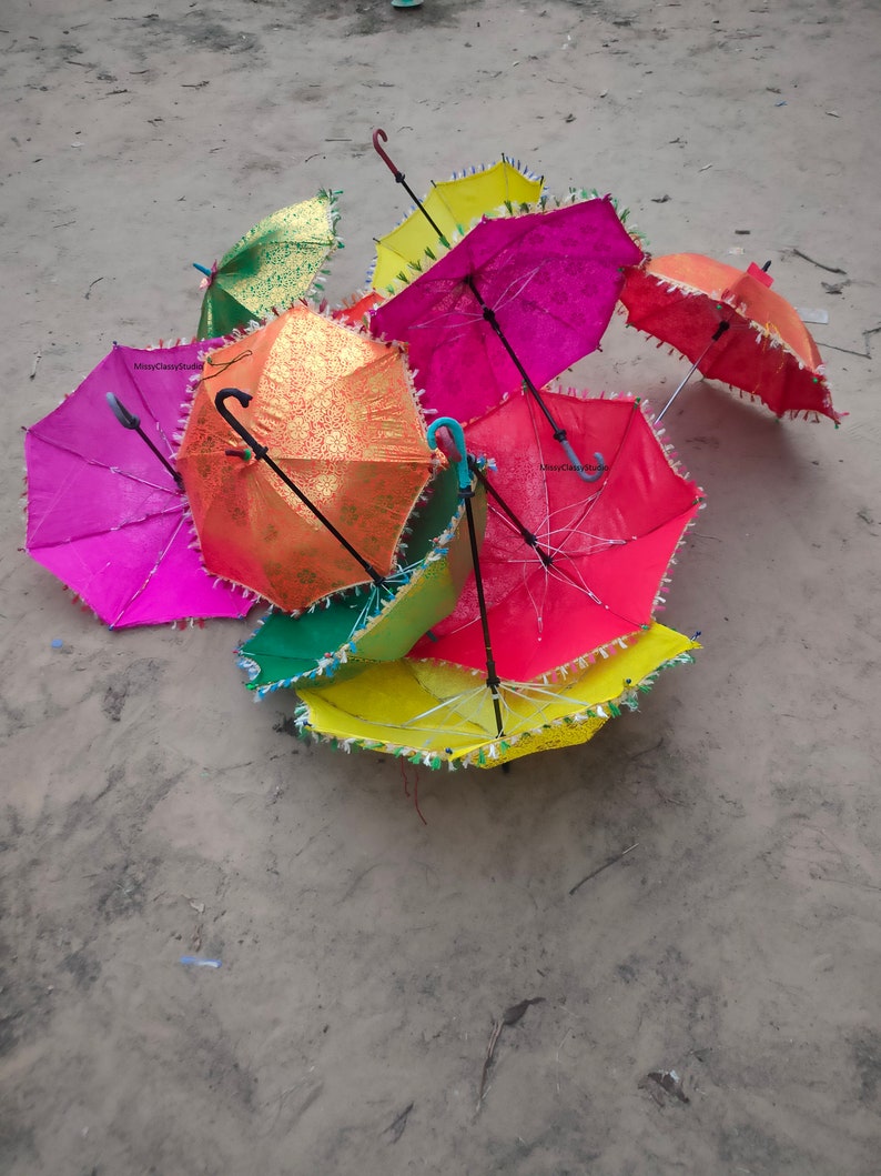 May include: A group of colorful umbrellas with fringe trim. The umbrellas are in various shades of pink, red, yellow, green, and orange. The umbrellas are lying on a sandy surface.