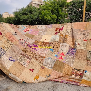 Peut inclure: Une courtepointe beige &agrave; motifs de patchwork avec des carr&eacute;s de diff&eacute;rentes couleurs marron, rose et violet. La courtepointe est drap&eacute;e sur un rebord, avec des arbres verts et un b&acirc;timent en arri&egrave;re-plan.