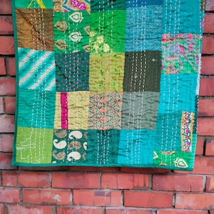 May include: A patchwork quilt made from various green, blue, and brown fabrics. The quilt is made up of squares of different sizes and patterns, and is stitched together with white thread. The quilt is hanging on a brick wall.