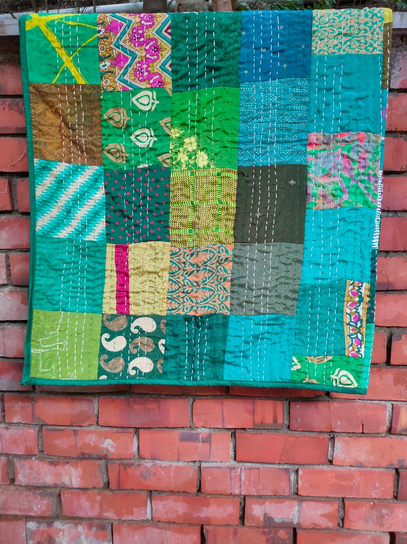 May include: A patchwork quilt made from various green, blue, and brown fabrics. The quilt is made up of squares of different sizes and patterns, and is stitched together with white thread. The quilt is hanging on a brick wall.
