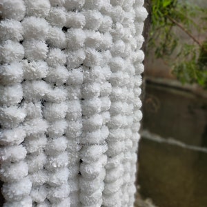May include: A white garland of artificial flowers, made of fluffy pom-poms, hangs against a wall. The garland is made of many small, white pom-poms, creating a soft and delicate look. The garland is perfect for adding a touch of elegance to any space.