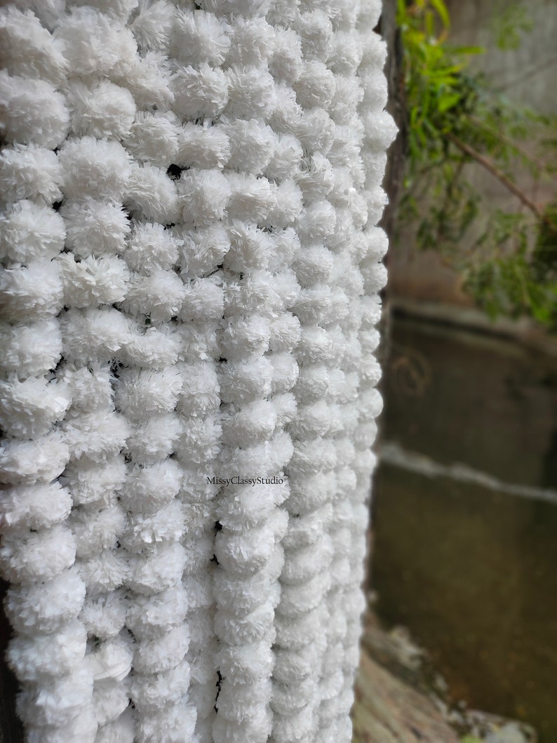 May include: A white garland of artificial flowers, made of fluffy pom-poms, hangs against a wall. The garland is made of many small, white pom-poms, creating a soft and delicate look. The garland is perfect for adding a touch of elegance to any space.