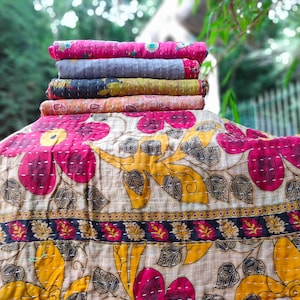 May include: A stack of colorful, rolled kantha quilts. The top quilt is pink, followed by gray, black, orange, and pink. The bottom quilt features a large floral design with pink and yellow flowers on a beige background.