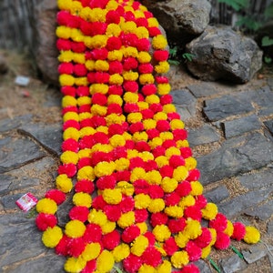 May include: A long garland of artificial marigold flowers in yellow and pink. The garland is made of multiple strands of flowers, each with a stem. The garland is lying on a stone path.