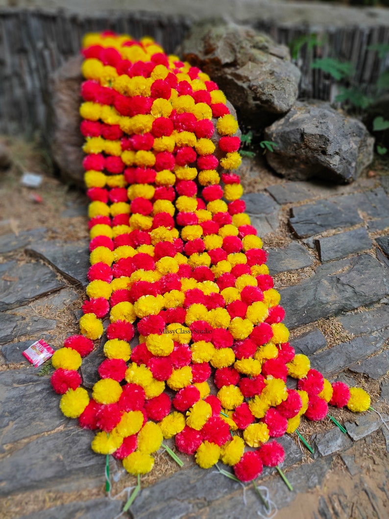 May include: A long garland of artificial marigold flowers in yellow and pink. The garland is made of multiple strands of flowers, each with a stem. The garland is lying on a stone path.
