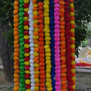 Può includere: Un'esposizione vibrante di ghirlande di fiori colorati. Le ghirlande sono disposte verticalmente, mostrando uno spettro di tonalità tra cui verde, giallo, arancione, bianco, blu, rosa e rosso. I fiori sembrano essere artificiali e sono infilati.