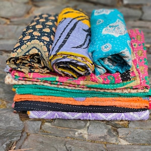 May include: A stack of colorful kantha quilts, a type of Indian patchwork quilt, with various patterns and designs. The quilts are folded and stacked on top of each other, creating a visually appealing display.