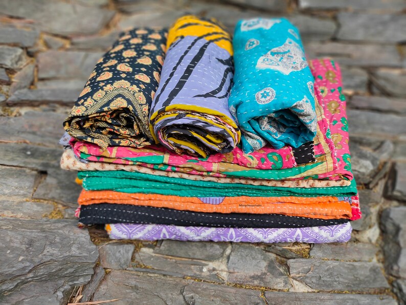 May include: A stack of colorful kantha quilts, a type of Indian patchwork quilt, with various patterns and designs. The quilts are folded and stacked on top of each other, creating a visually appealing display.