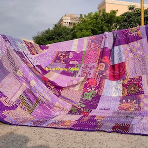 Peut inclure: Une courtepointe patchwork dans les tons de violet, lavande et magenta, avec une bordure violet fonc&eacute;. La courtepointe est faite de divers motifs de tissu, y compris des motifs floraux et g&eacute;om&eacute;triques. La courtepointe est expos&eacute;e &agrave; l'ext&eacute;rieur, drap&eacute;e sur une barre m&eacute;tallique.