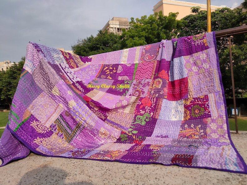Peut inclure: Une courtepointe patchwork dans les tons de violet, lavande et magenta, avec une bordure violet fonc&eacute;. La courtepointe est faite de divers motifs de tissu, y compris des motifs floraux et g&eacute;om&eacute;triques. La courtepointe est expos&eacute;e &agrave; l'ext&eacute;rieur, drap&eacute;e sur une barre m&eacute;tallique.