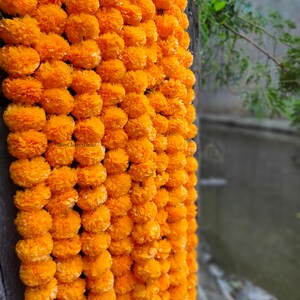 May include: A long garland of bright orange marigold flowers, strung together and hanging from a wooden surface. The garland is made of paper flowers and is a popular decoration for Indian weddings and festivals.