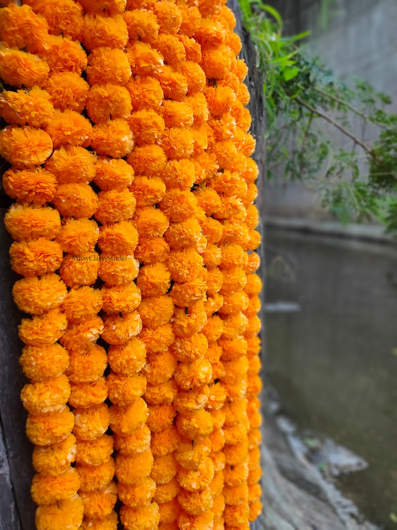 May include: A long garland of bright orange marigold flowers, strung together and hanging from a wooden surface. The garland is made of paper flowers and is a popular decoration for Indian weddings and festivals.