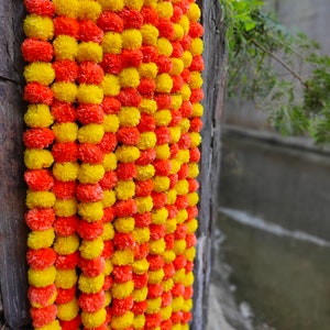 May include: A garland of yellow and orange marigold flowers, strung together in a long strand. The garland is hanging on a wooden surface.