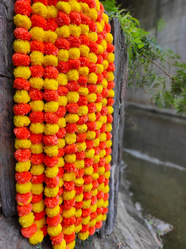 May include: A garland of yellow and orange marigold flowers, strung together in a long strand. The garland is hanging on a wooden surface.
