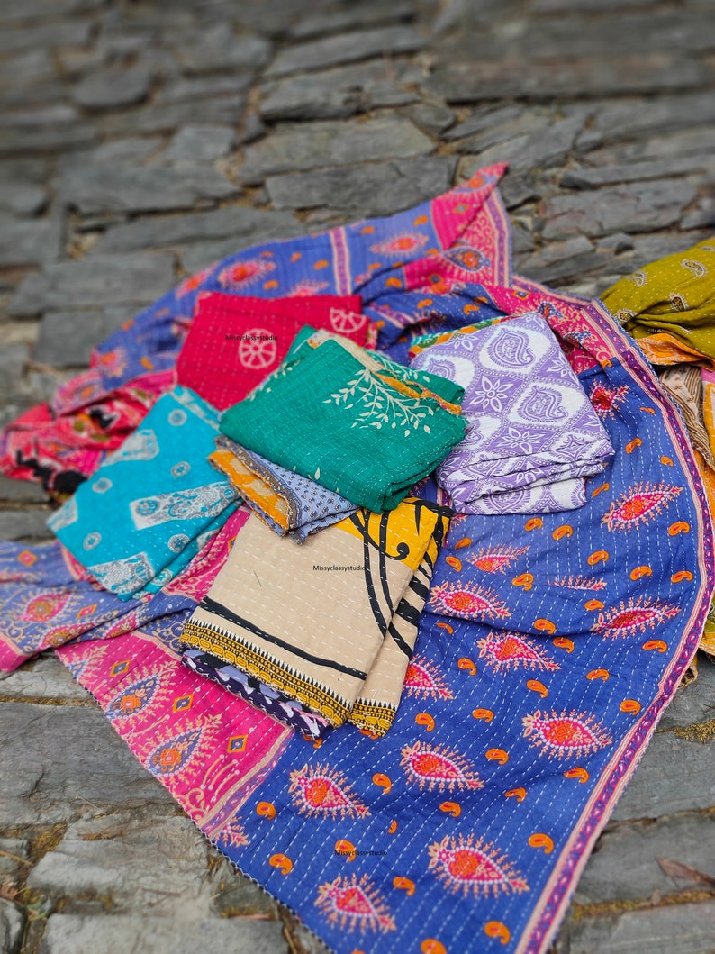 May include: A stack of colorful kantha quilts with various patterns, including paisley, floral, and geometric designs. The quilts are made of recycled saris and are perfect for adding a touch of bohemian style to any home.