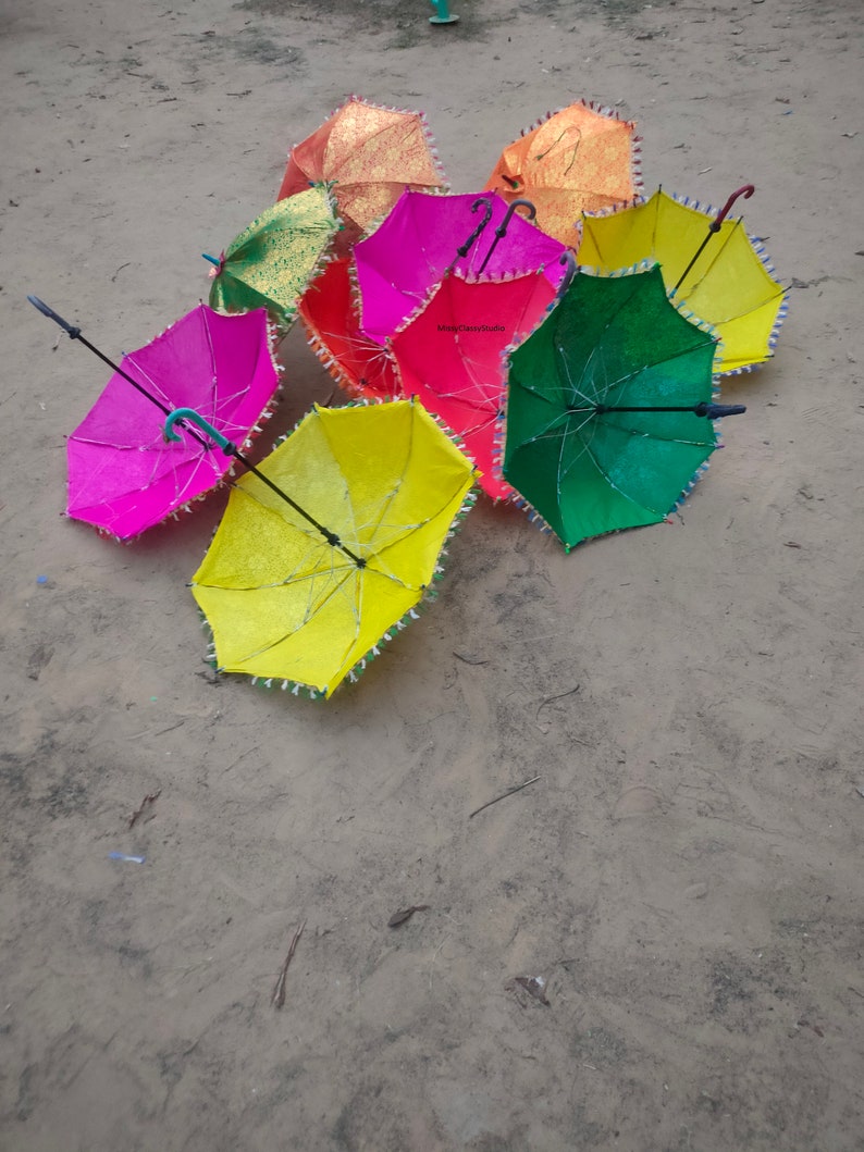 May include: A group of colorful umbrellas with fringed edges. The umbrellas are in various shades of pink, green, yellow, and orange. The umbrellas are arranged in a circle on a sandy surface.