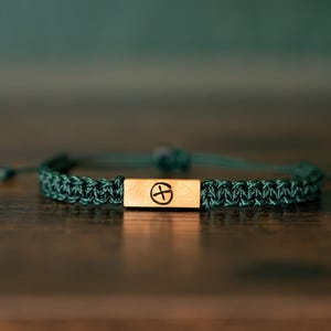 Puede incluir: Una pulsera de cordón trenzado verde oscuro con una barra metálica rectangular de tono dorado en el centro. La barra de metal está grabada con un símbolo circular que contiene una cruz. La pulsera tiene extremos de cordón ajustables.