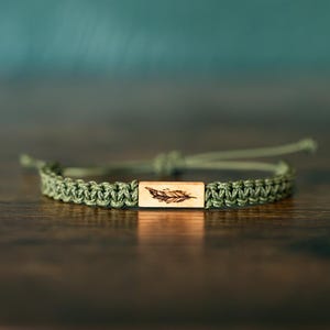 May include: An olive green cord bracelet featuring a rectangular wooden centerpiece. The wooden charm is engraved with a detailed feather illustration. The bracelet is constructed with a braided cord design and includes adjustable pull strings for sizing.