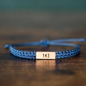 May include: A blue cord bracelet featuring a rectangular metallic bar centred on the band. The bar is engraved with the number 143 in a dark font. The bracelet includes adjustable cord ends with knots, providing a secure fit for the wearer.