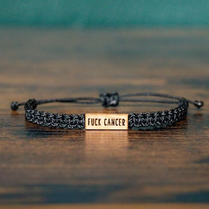May include: A black cord bracelet featuring a rectangular wooden charm centered on the band. The wooden piece is engraved with the text FUCK CANCER in a bold, black, sans-serif font. The bracelet has an adjustable knot closure system.